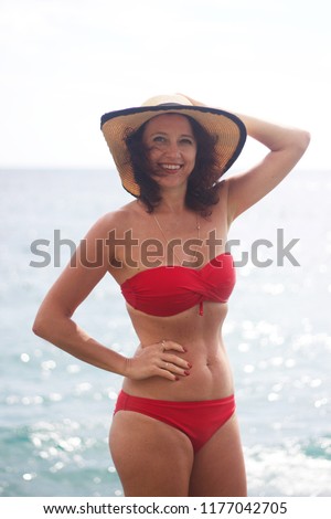 An awesome, smiling, curly-haired woman in red swimwear and beach hat is standing on the pier in the backdrop of the sea  Royalty-Free Stock Photo #1177042705