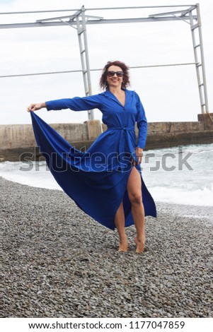 Lovely , curly-haired woman in blue dress is standing on the pebble beach
