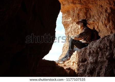 MAN WITH BACKPACK AND AUSTRALIAN HAT SITTING IN A CAVE LOOKING AT THE LANDSCAPE