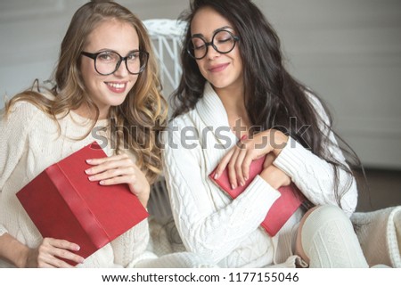 Young friends in glasses and knitted clothes are sitting on a chair with books. Girls enjoy communication and reading