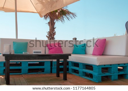 Reserved beach table with sofas from euro pallets and bright soft pillows. A reserved table under the beach umbrella. Large leather sofas of euro pallets with green and pink cushions. Beach style. Royalty-Free Stock Photo #1177164805