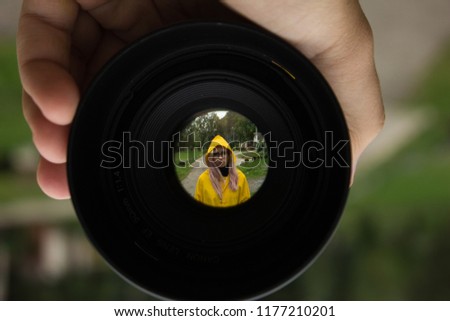 soft focus sad girl portrait in yellow raincoat in creative foreshortening through camera lens with circle black frame, unfocused blurred background 