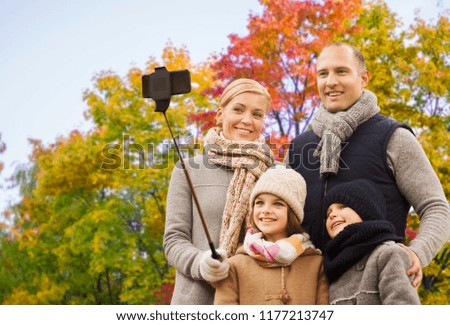 family, technology and people concept - happy mother, father, daughter and son taking picture by smartphone on selfie stick over autumn park background