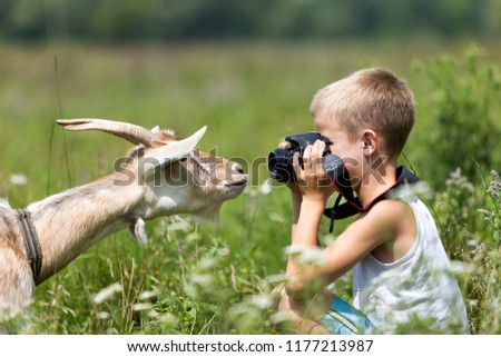 Profile portrait of young blond cute handsome child boy taking picture of funny curious goat looking straight in camera on bright sunny summer day on blurred light green grassy copyspace background.
