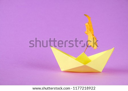 Yellow paper boat from origami with an autumn leaf on a purple background.
