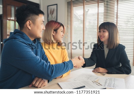 Happy young Asian couple and realtor agent. Cheerful young man signing some documents and handshaking with broker while sitting at desk. Signing good condition contract.