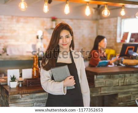 Portrait of Asian girl waitress holding menu wearing apron and standing in coffee shop. Royalty-Free Stock Photo #1177352653