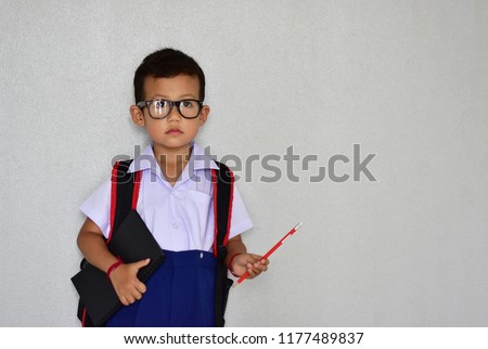 Back to school, little nerdy Asian kindergarten boy in school uniform wearing glasses carrying a bag and a notebook ready to go to school on light grey background, first day to school