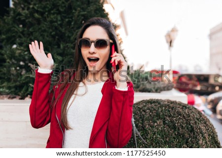 Beautiful young woman with long brown hair expressing amazement while talking on phone. Outdoor photo of cheerful surprised girl posing with smartphone.