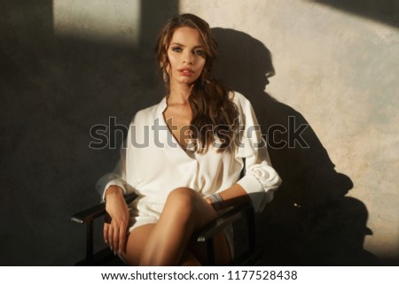 Portrait of yong woman in white blouse and shorts sitting at black high chair. Fashion photo with hard sunlight with dark shadows