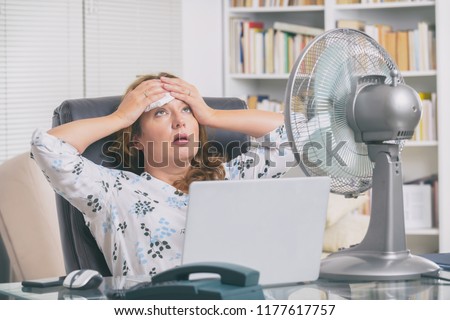 Woman suffers from heat while working in the office and tries to cool off by the fan Royalty-Free Stock Photo #1177617757