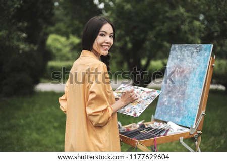 Artist painting easel outdoor. Authentic creative teacher painting with palette watercolor, oil, acril paints palette and brush morning sunlight. View of nature and garden exterior, outdoors workshop
