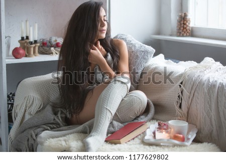 A young blonde with a book in her hands sits on the couch. A girl in a knitted sweater enjoys reading in a cozy room