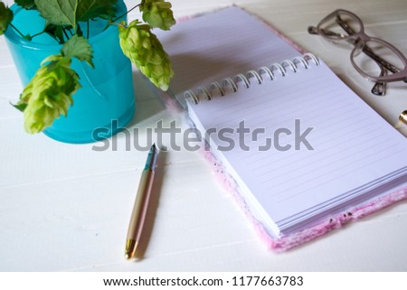 The opened notepad, pen, white candle, glasses and branches of hops as decoration on a white wooden table. Desktop still life with space for text.
