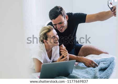 joy and happy beautiful caucasian couple at home in the bedroom on the bed using computer and taking a selfie picture with the smartphone. laugh and happiness for boy and girl in our life. cheerful 