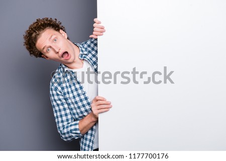 Stylish trendy nice handsome shocked funky positive young guy with wavy hair in casual checkered shirt, hiding, holding big wide white promo board, blank empty place. Isolated over grey background