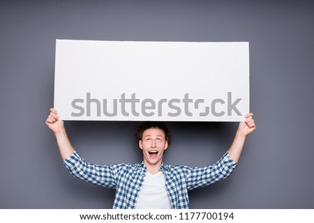 Stylish trendy attractive nice handsome cheerful funky positive young man with wavy hair in casual checkered shirt, holding big wide white promo board, copy-space. Isolated over grey background