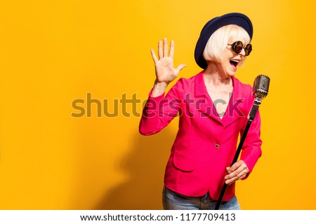 Portrait of charming modern grandmother holds up the microphone stand and sings a raised palm top isolated on vivid yellow background with copy space for text Royalty-Free Stock Photo #1177709413