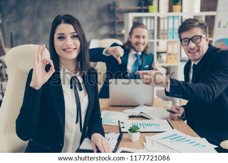 Three business people, elegant classic nice lovely lady boss showing ok sign, two partners showing thumb down and pointing to girl, wearing suits, jackets at work place, work station