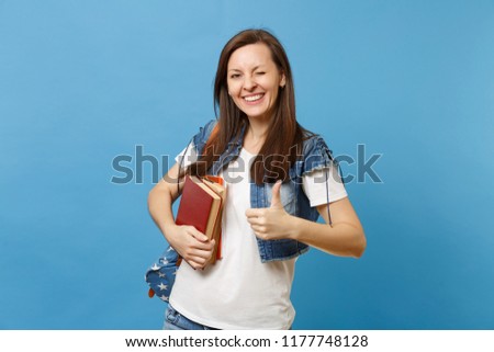 Portrait of young funny cute woman student in denim clothes with backpack blinking showing thumb up, hold school books isolated on blue background. Education in high school university college concept