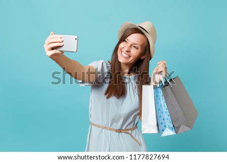 Portrait smiling woman in summer dress straw hat holding packages bags with purchases after shopping doing selfie shot on mobile phone isolated on blue pastel background. Copy space for advertisement