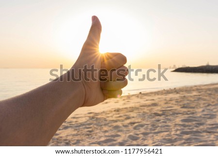 Male hand doing thumbs up or good gesture with sunrise background at the beach. Sunburst effect on hand.