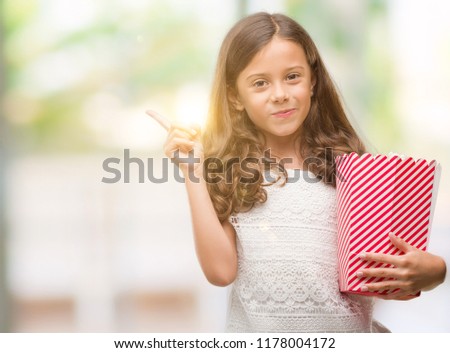 Brunette hispanic girl eating popcorn very happy pointing with hand and finger to the side