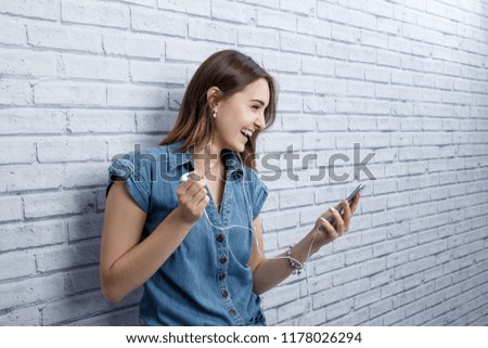 Young girl student teenager listening to music wearing earphones holding smart phone standing near the grey brick wall smiling laughing