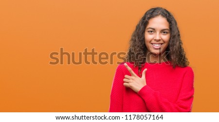 Young hispanic woman wearing red sweater cheerful with a smile of face pointing with hand and finger up to the side with happy and natural expression on face looking at the camera.