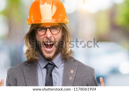 Young handsome architec man with long hair wearing safety helmet over isolated background shouting with crazy expression doing rock symbol with hands up. Music star. Heavy concept.