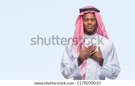 Young arabic african man wearing traditional keffiyeh over isolated background smiling with hands on chest with closed eyes and grateful gesture on face. Health concept.