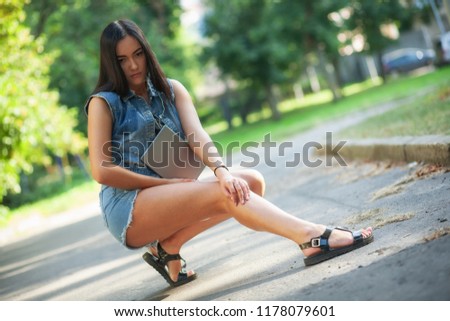 Young pretty brunette girl in denim shorts with the tablet is sitting on the road and listening to music on the street in summer.