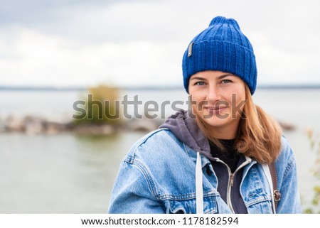 Portrait of outdoor atmospheric lifestyle photo of young beautiful  darkhaired woman  in knitting hat, in a denim jacket and black trousers against the background sea  in sunny autumn day  