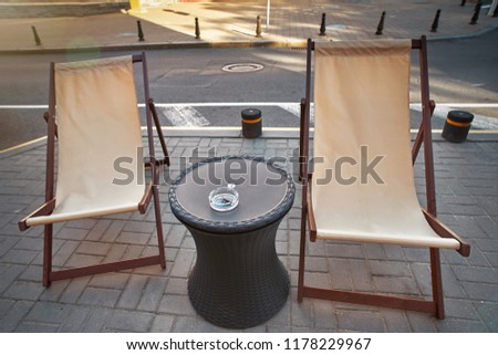 Cafe corner with deck chairs and round serving table of the summer cafe-restaurant near the street 
Lounge cafe terrace with chairs and coffee table with ashtray on it. Empty table of summer terrace