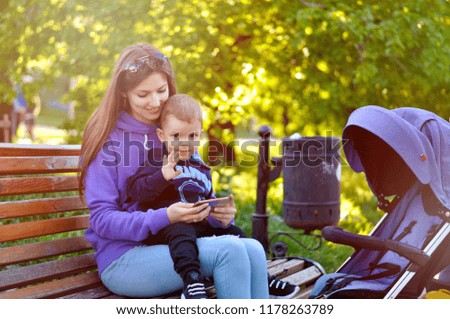 A young beautiful mother sits with her little son on a bench in the park and looks into the phone. They talk on video calling with the dad sailor.