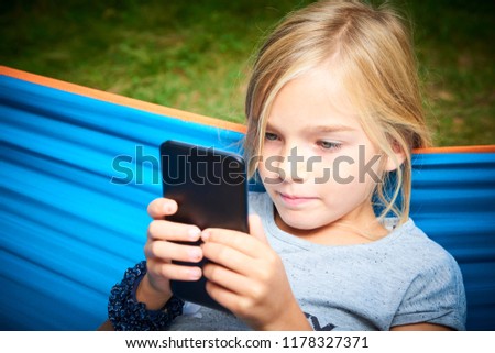 Child blond girl playing with a smart phone while lying on hammock outdoors 
