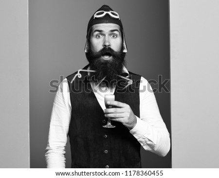 handsome bearded pilot or aviator man with long beard and mustache on surprised face holding glass of alcoholic shot in vintage suede leather waistcoat with hat and glasses on studio background.