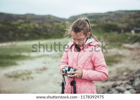 Bespectacled girl in a pink jacket takes pictures on the dirt road
