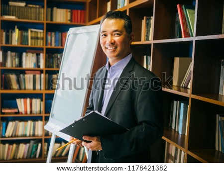 Nice portrait of asian stylish man wearing suit. Man smiling and looking at camera. Big bookshelf as a background.