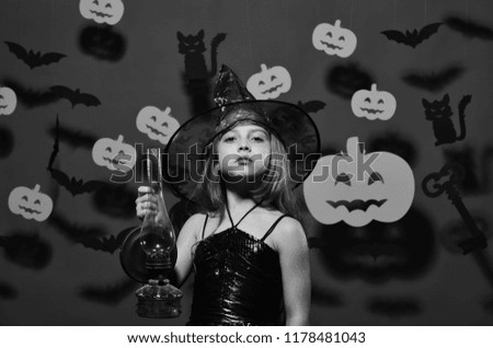 Halloween party and decorations concept. Little witch wearing black hat. Girl with proud face on pink background with bats and pumpkins decor. Kid in spooky witches costume holds old gas lamp