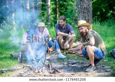 Hipster roasting sausage while friends speaking sharing impression and watching photos on camera. Tourists having snack time with roasted over fire food. Friends group tourist relaxing near bonfire.