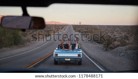 Group Of Friends On Road Trip Driving Classic Convertible Car Viewed Through Windshield Of Following Vehicle Royalty-Free Stock Photo #1178488171