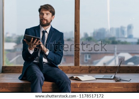 a man in a suit sits on a window sill with a tablet in his hands                         