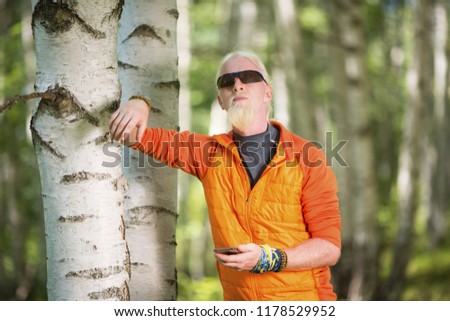 Young man with a blonde hair and beard holding smartphone and leaning on a tree in nature 
