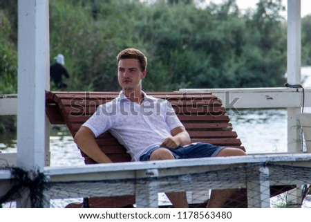 A young attractive man in a white shirt sits on a floating restaurant in the background of a pond