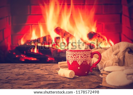 Mug of hot chocolate or coffee with marshmallows in a red mug on vintage wood table in front of Fireplace as a background. Christmas or winter warming drink.