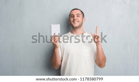 Young caucasian man over grey grunge wall showing notebook surprised with an idea or question pointing finger with happy face, number one