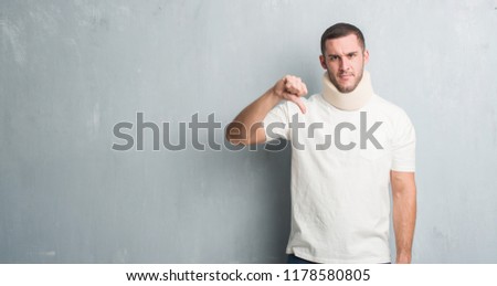 Young caucasian man injured over grey grunge wall wearing neck collar with angry face, negative sign showing dislike with thumbs down, rejection concept