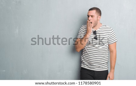 Young caucasian man over grey grunge wall bored yawning tired covering mouth with hand. Restless and sleepiness.