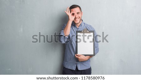 Young caucasian man over grey grunge wall holding clipboard with happy face smiling doing ok sign with hand on eye looking through fingers
